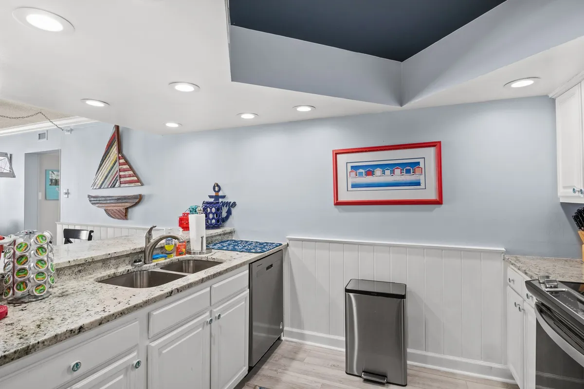 A cheerful kitchen scene with granite countertops, colorful wall art, a decorative sailboat, and a blue kitchen mat. The space feels inviting and well-equipped, ideal for both casual breakfasts and festive gatherings.