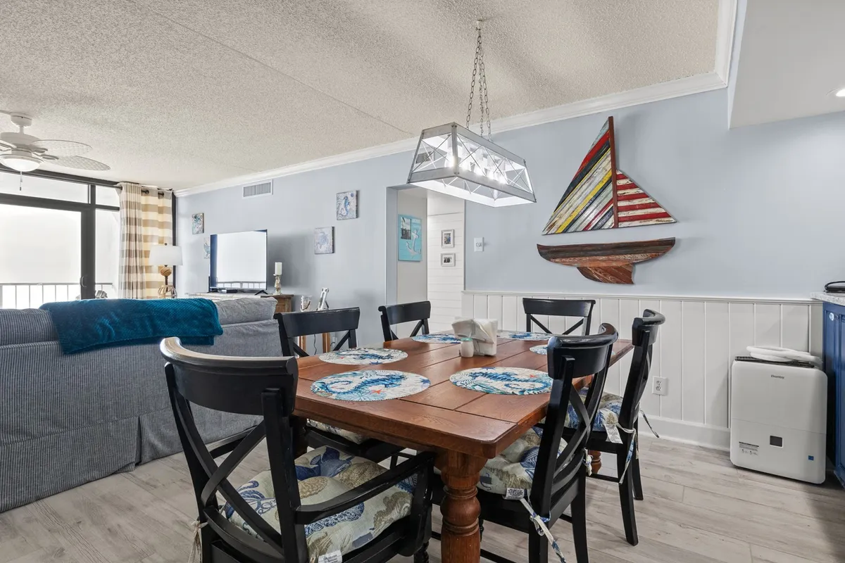 A sunny dining area featuring a welcoming table set beneath a chandelier, sailboat wall art, and an adjacent living room couch. The space is bathed in natural light, creating a warm atmosphere perfect for group meals or intimate dinners.