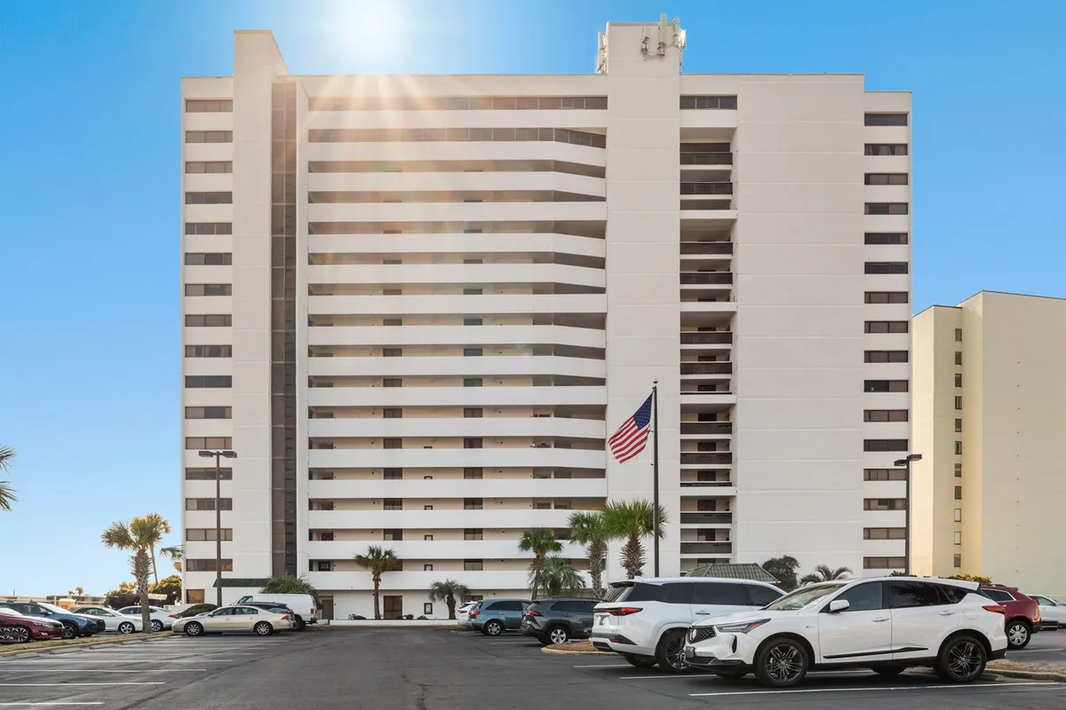 A tall oceanfront condominium building is surrounded by mature palm trees, with the American flag gently waving in the foreground. The parking lot is visible, indicating easy access for guests, and the welcoming facade hints at the convenience and comfort of beachfront living. The ocean is glimpsed just beyond the grounds, creating a seamless connection to the sand and surf.