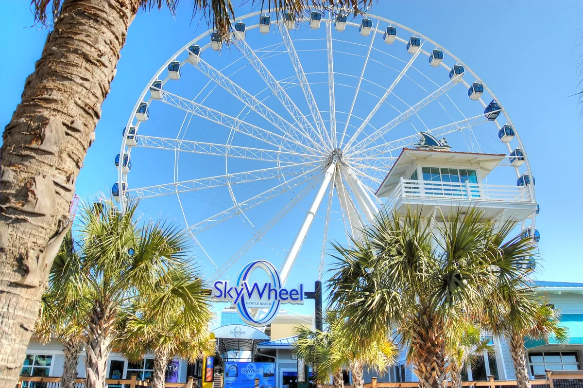 The iconic Myrtle Beach SkyWheel rises above palm trees and a bustling park area, with blue skies and the building facade in the background. The scene is vibrant, with hints of activity and adventure, alluding to the nearby attractions and family fun highlighted in the section content.