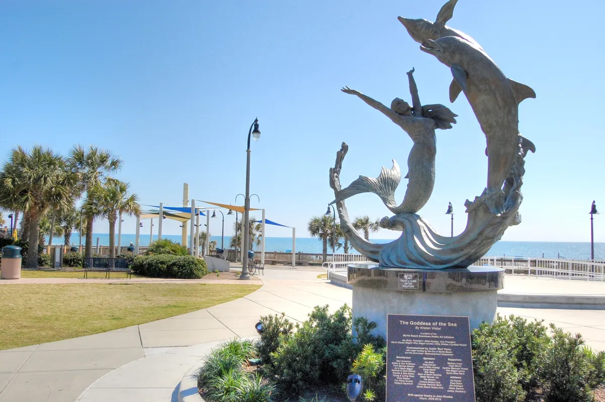 A picturesque walkway lined with palm trees leading to a statue of the Goddess of the Sea, with expansive ocean views and a relaxed, open-air vibe.