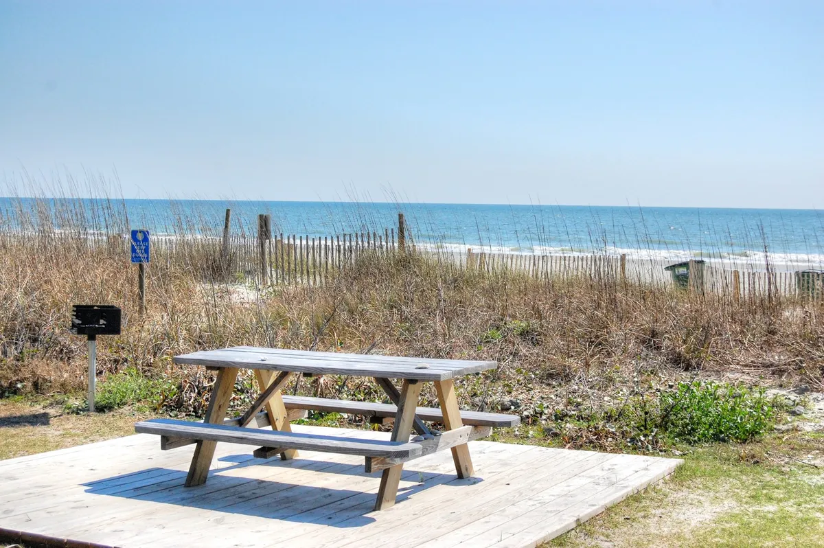 A serene picnic area featuring a wooden picnic table set against a backdrop of dune grass, a wooden fence, and sweeping ocean views. The setting invites guests to enjoy alfresco meals while soaking in the peaceful coastal ambiance.