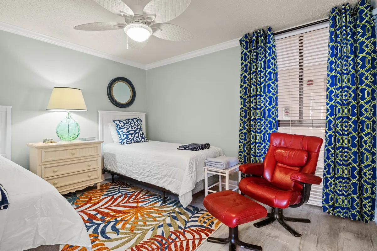 A vibrant living area with a colorful area rug, a red chair, decorative lamp, and textured curtains. The space feels playful and flexible, inviting guests to relax, read, or socialize in comfort.