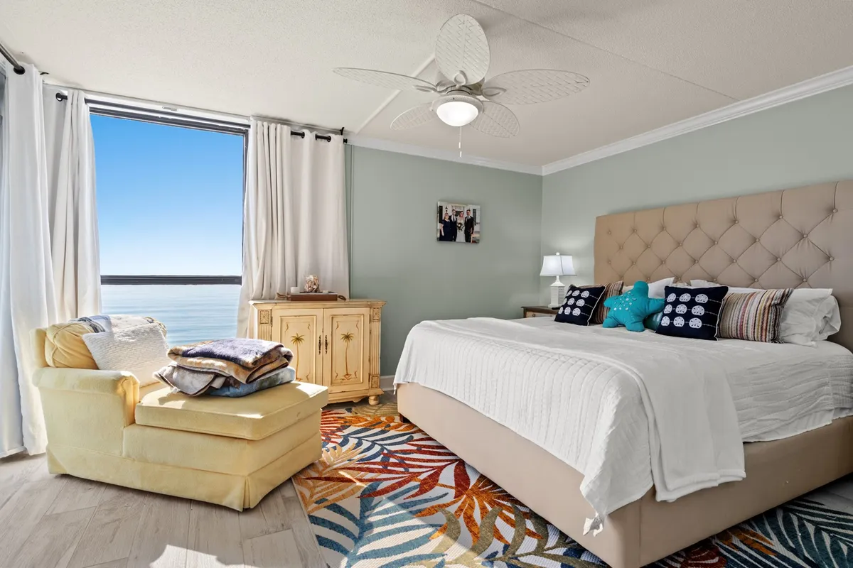 A serene master bedroom featuring a tufted King bed, large window overlooking scenic water views, a colorful area rug, and a comfortable armchair. The room is bathed in natural light and styled with calming décor, creating an inviting retreat for relaxation.