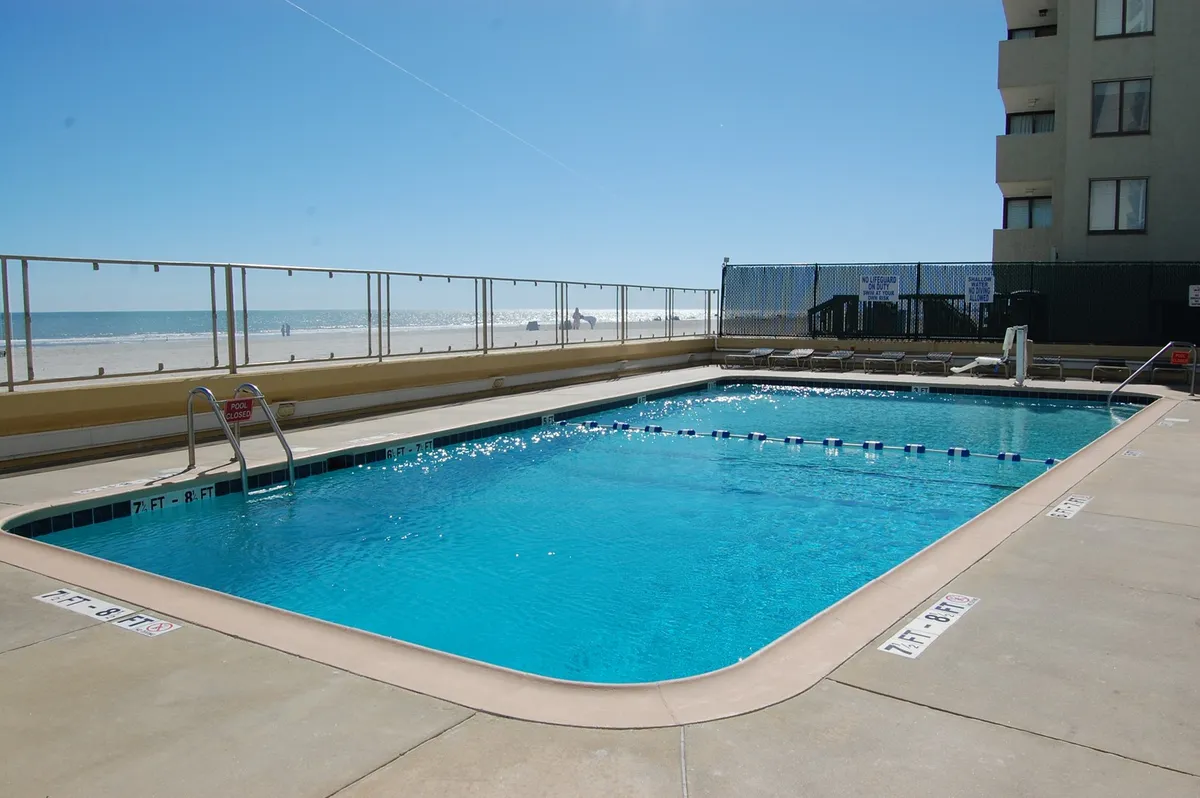 This image presents a vibrant, oceanfront swimming pool surrounded by deck chairs, with the sparkling Atlantic Ocean visible just beyond. The scene is lively, embodying the leisurely, resort-style experience with ample space for socializing, swimming, and sunbathing.