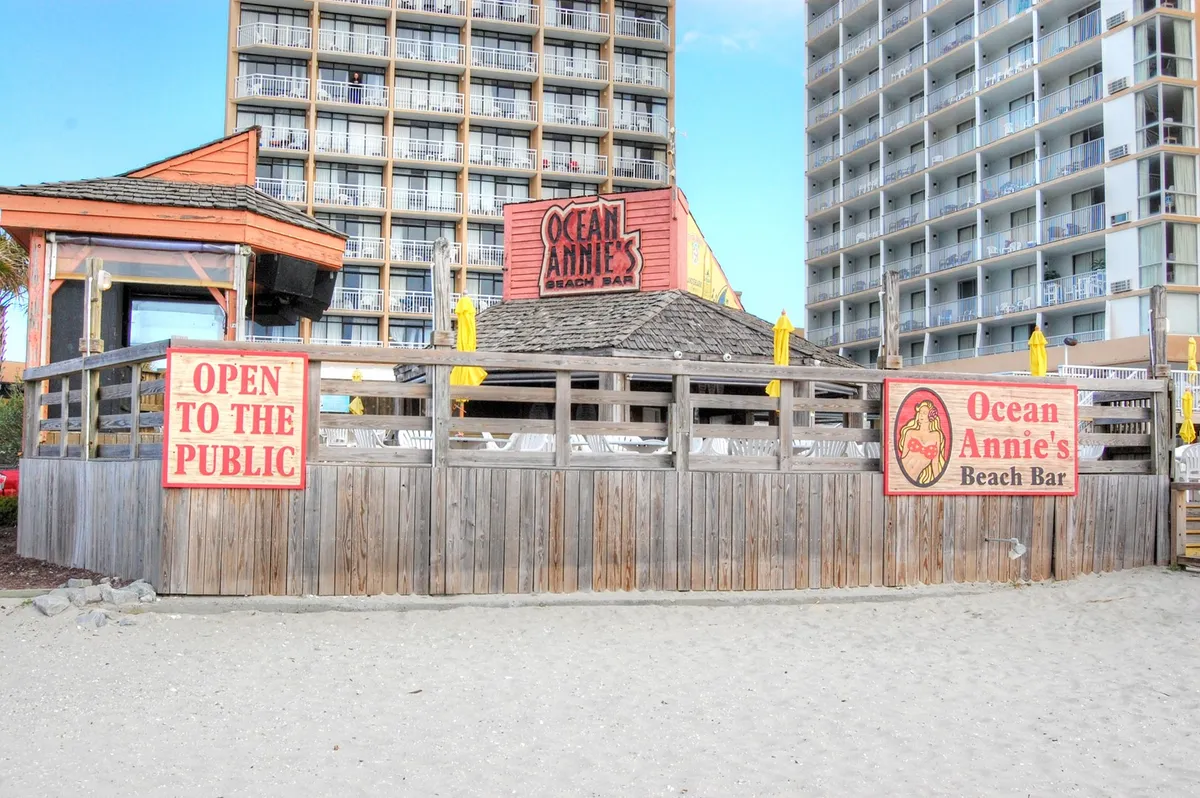 A lively beachfront bar featuring the 'Ocean Annie's Beach Bar' sign, vibrant yellow umbrellas, and a wooden deck overlooking the sand, capturing a festive coastal ambiance.