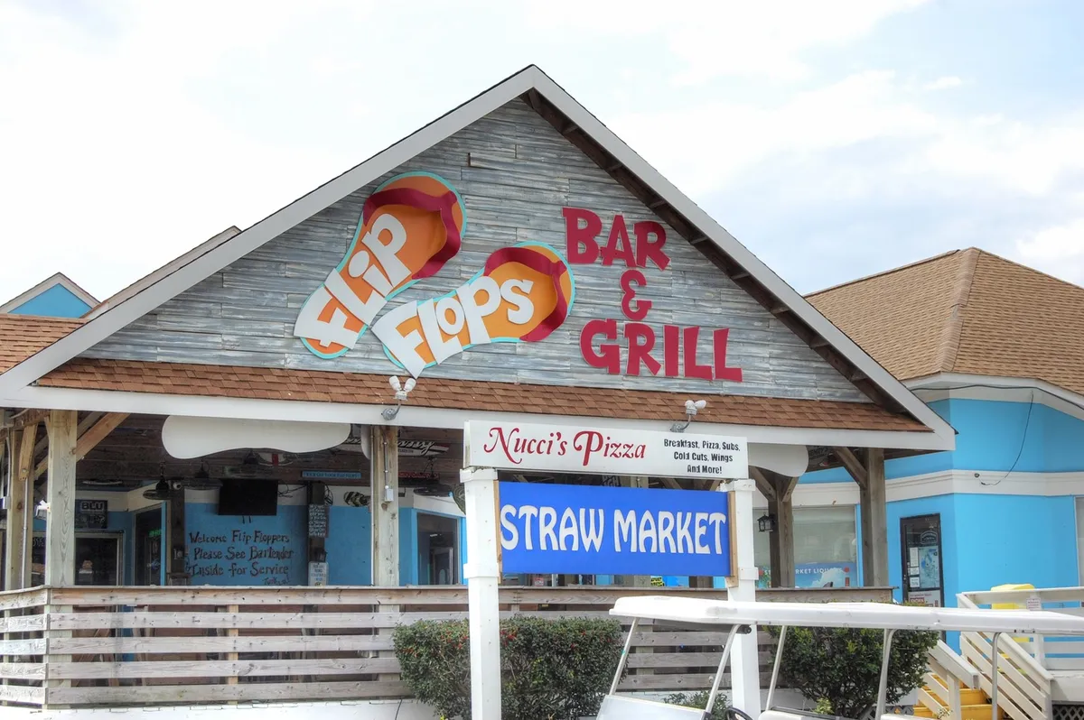 An exterior view featuring signage for 'Flip Flops' Bar & Grill and the 'Straw Market,' with outdoor seating visible and a relaxed, social atmosphere. The image suggests opportunities for dining, live music, and community engagement in a coastal setting.