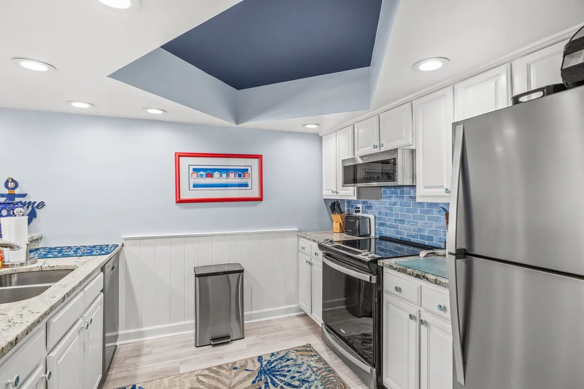 This image features a modern kitchen with a granite countertop, blue backsplash, colorful artwork, and stylish appliances. The space is bright and inviting, with ample counter space for meal prep, and visually appealing kitchen decor that enhances the contemporary coastal vibe.