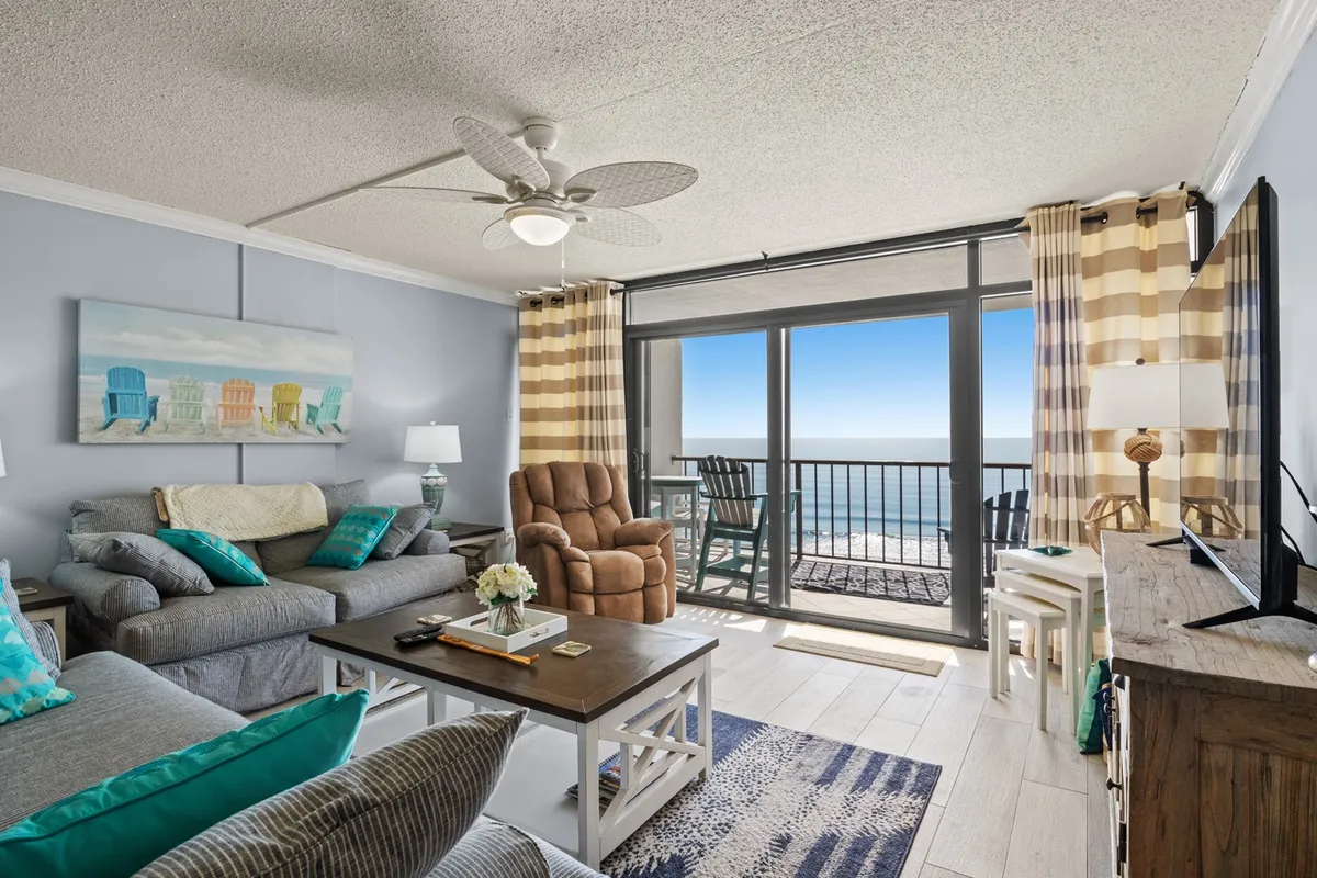 The image showcases a stylish condo living area with a plush sofa adorned with vibrant cushions, a wooden coffee table with artistic decor, and expansive floor-to-ceiling windows framing sweeping ocean views. Through the windows, a cozy balcony with a rocking chair invites guests to relax while overlooking the Atlantic, highlighting the serene coastal ambiance and luxurious comfort of the space.