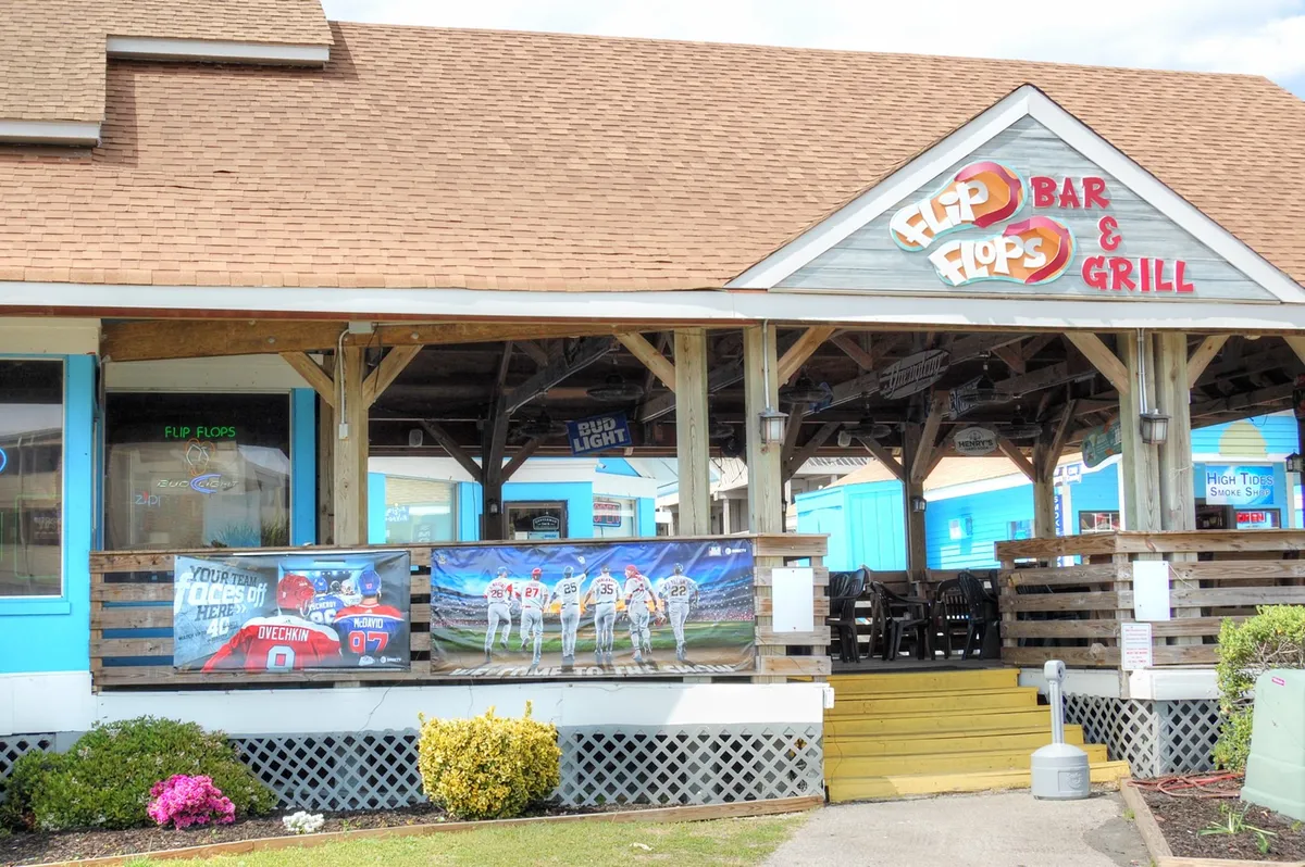 A vibrant exterior shot showcasing the entrance to Flip Flops Bar & Grill, with colorful sports banners and a bustling outdoor seating area. People can be seen dining, socializing, and enjoying the coastal breeze.