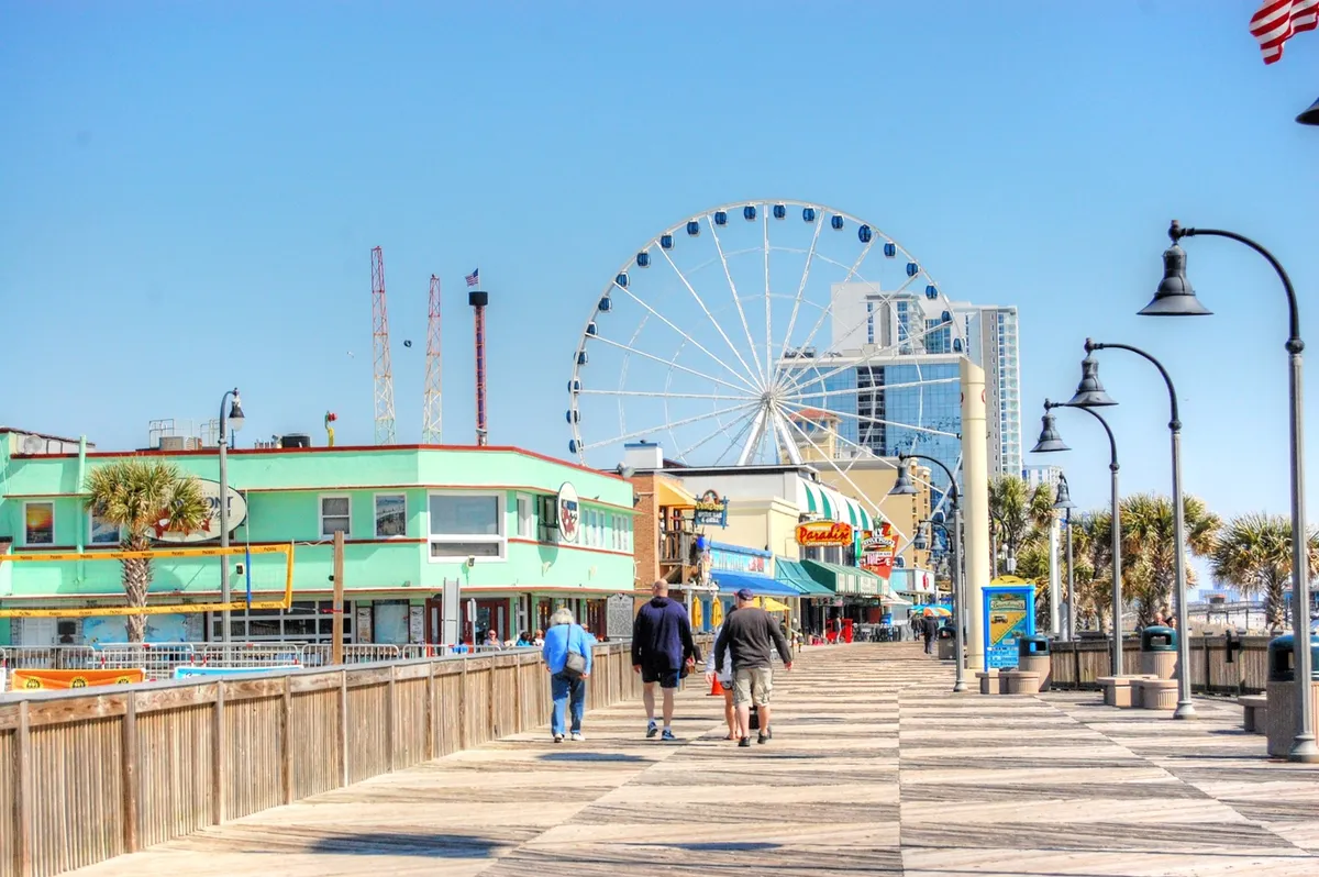 This lively photo shows a bustling boardwalk scene complete with a ferris wheel, colorful buildings, and people strolling and enjoying the vibrant atmosphere. It captures the excitement of local attractions and the joy of shared outdoor experiences.